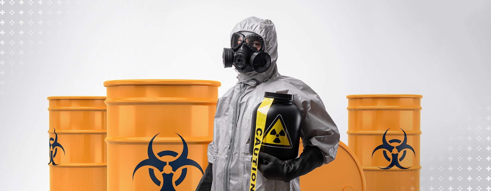 Imagem mostra uma pessoa vestindo um traje de proteção que a cobre completamente, enquanto segura um pote preto com o símbolo de radioatividade. Ao fundo, estão três barris laranjas também com o símbolo de radioatividade