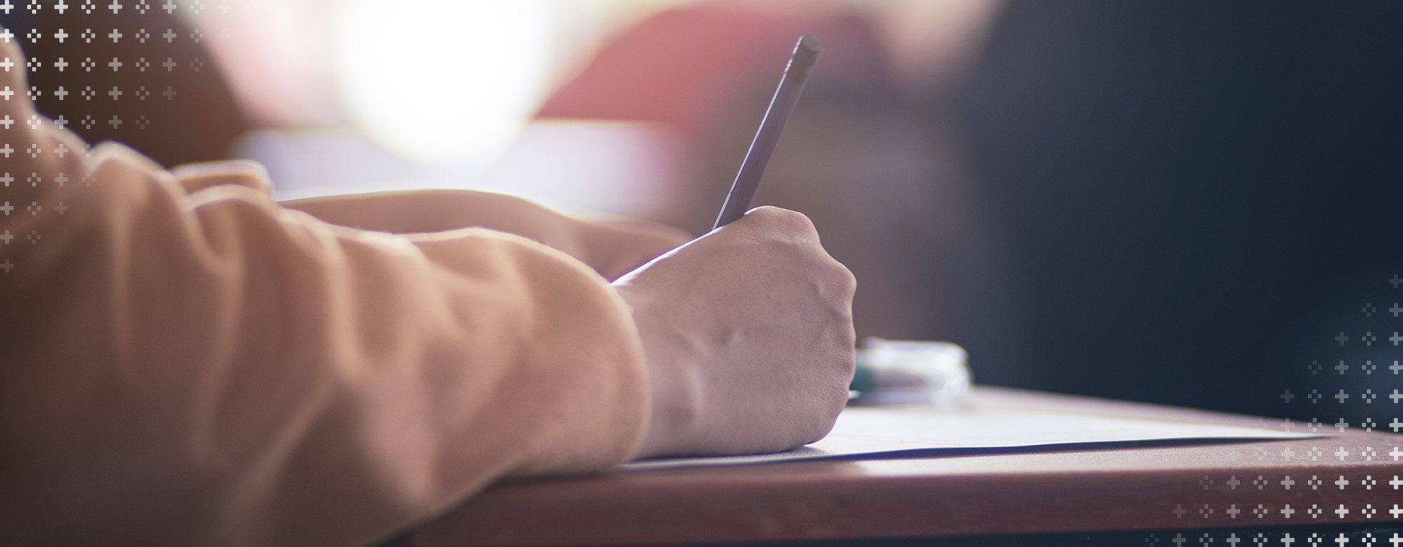 imagem de uma mão posicionada sobre uma mesa, segurando uma caneta, escrevendo uma redação
